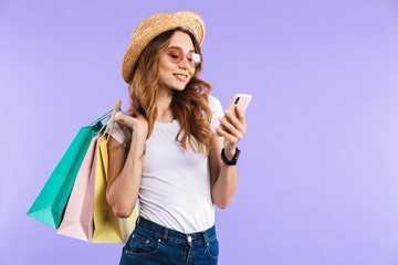 Wall Mural - Happy cute woman isolated over purple wall background holding shopping bags using mobile phone.