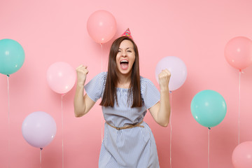 Portrait of attractive joyful young woman in birthday hat blue dress doing winner gesture screming on pink background with colorful air baloons. Birthday holiday party, people sincere emotion concept.