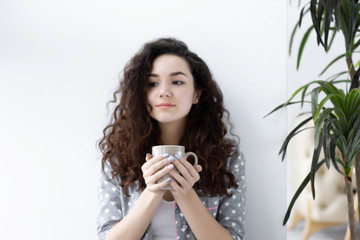 Wall Mural - Portrait of young pretty girl woman female wearing pajamas sleepwear with white hearts sitting near white wall drinking cup of coffee tea and smiling in the morning. Morning wake up dreaming concept