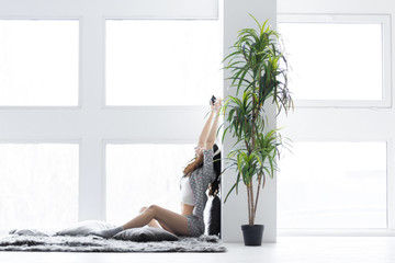 Wall Mural - Young girl woman female adult wearing grey pajamas white top and socks sitting on blanket near the large windows, relaxing and listening to music in the light interior room with plant. Morning concept