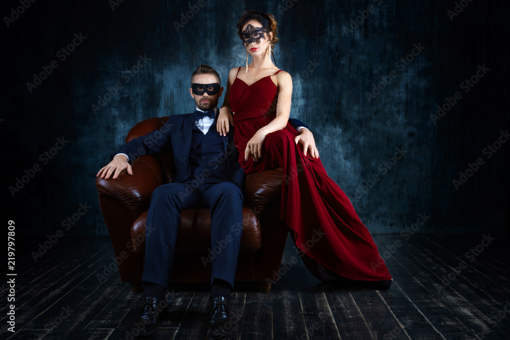 Rich couple man male woman female in love sitting in leather sofa in  expensive suit and red evening dress and black carnaval mask. Sex, tempts,  harassment, sexism, seduction issues Stock Photo |