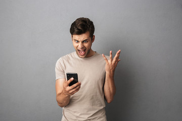 Canvas Print - Angry man 20s with short dark hair screaming in rage while holding smartphone, isolated over gray background