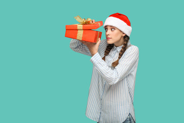 Wall Mural - New year concept. cunning beautiful girl in striped light blue shirt in red christmas cap standing holding red gift box, unboxing and looking inside. indoor, studio shot isolated on green background.