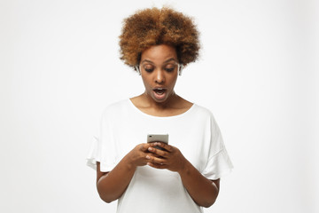 Beautiful african american woman isolated on gray background, looking to screen of smartphone with mouth round because of deep astonishment and shock of great news