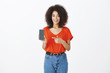 © Cookie Studio - Portrait of good-looking african-american shop assistant in trendy red t-shirt and jeans, showing smartphone and pointing at device with index finger, suggesting cusomet to by brand new cool phone