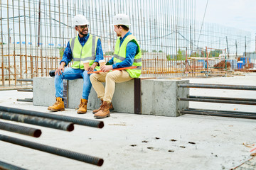 Full length portrait of two workers wearing hardhats  chatting while enjoying coffee break on construction site