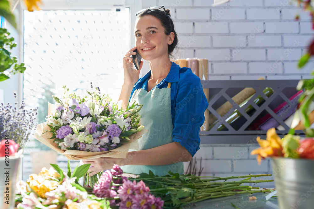 Young smiling florist holding flowers and connecting on mobile phone near workplace - obrazy, fototapety, plakaty 