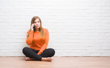 Wall Mural - Young adult woman sitting on the floor over white brick wall calling using smartphone with a confident expression on smart face thinking serious