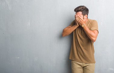 Handsome young man over grey grunge wall with sad expression covering face with hands while crying. Depression concept.