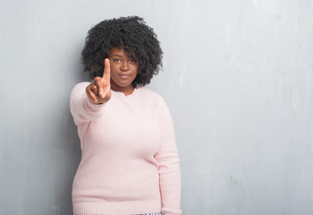 Poster - Young african american plus size woman over grey grunge wall wearing winter sweater showing and pointing up with finger number one while smiling confident and happy.