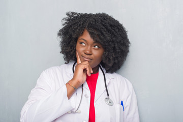 Sticker - Young african american doctor woman over grey grunge wall with hand on chin thinking about question, pensive expression. Smiling with thoughtful face. Doubt concept.