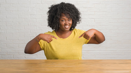 Wall Mural - Young african american woman sitting on the table at home looking confident with smile on face, pointing oneself with fingers proud and happy.