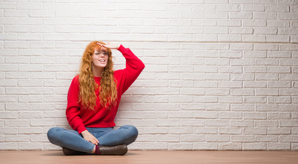 Poster - Young redhead woman sitting over brick wall very happy and smiling looking far away with hand over head. Searching concept.