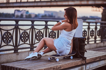 Wall Mural - Back view of a skater girl dressed in shorts and t-shirt sitting on the embankment during beautiful sunset.