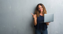 Middle Age Hispanic Woman Standing Over Grey Grunge Wall Using Laptop Very Happy Pointing With Hand And Finger To The Side