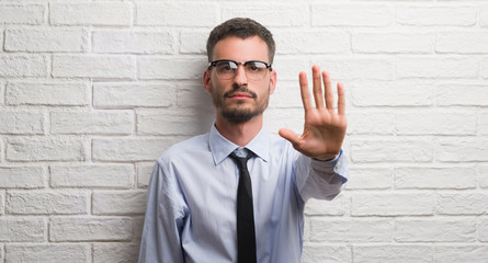 Poster - Young adult business man standing over white brick wall with open hand doing stop sign with serious and confident expression, defense gesture