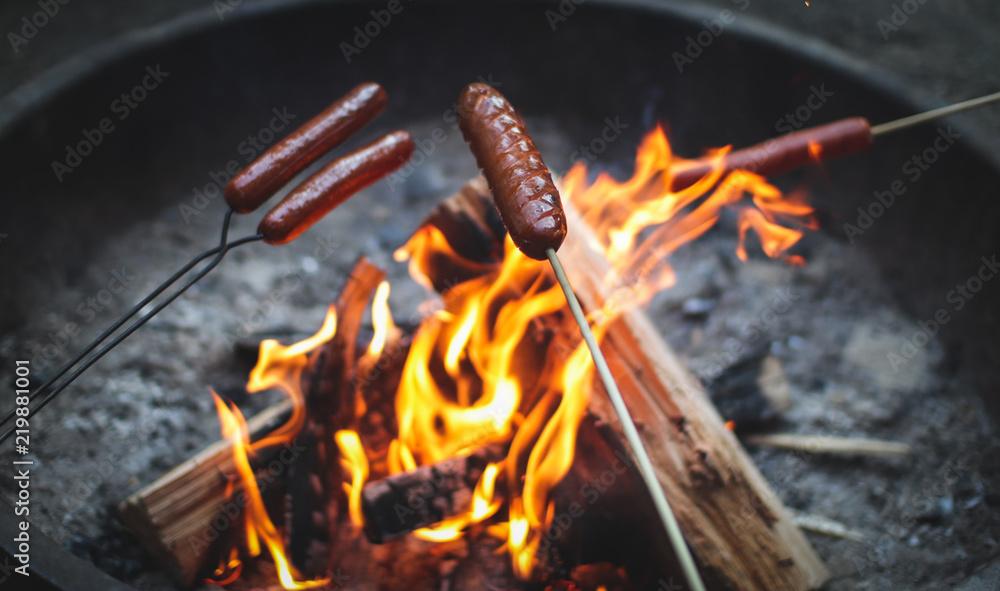 hotdogs roasting on sticks over flames in a campfire - obrazy, fototapety, plakaty 