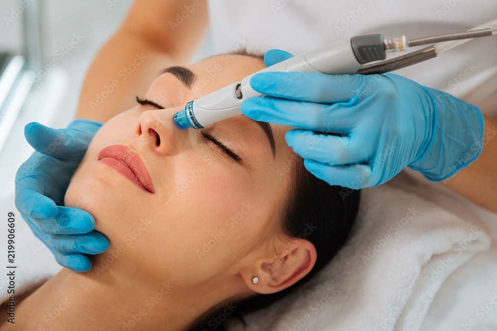 Facial procedure. Delighted nice woman lying on the medical bed with her eyes closed while having hydrafacial - obrazy, fototapety, plakaty 