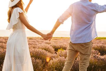 Canvas Print - Image from back of caucasian couple man and woman holding hands, while walking outdoor in summer lavender field