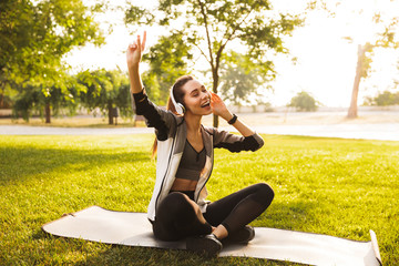 Wall Mural - Photo of happy sporty woman 20s in sportswear listening to music and singing, while sitting on exercise mat in green park