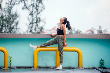 Portrait of a young, attractive and cool Asian millennial teenager girl leaning against a wall with a city in the backdrop. She is tanned, athletic and wearing trendy but simple street clothing.