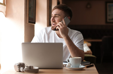 Canvas Print - Young freelancer talking on mobile phone while working on laptop in cafe