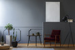 © Photographee.eu - Real photo of a red armchair standing between a black lamp and a table in a grey living room with striped rug, plants and empty poster mockup on a shelf