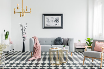 Pink blanket on grey sofa in living room interior with poster and patterned armchair. Real photo