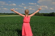 Młoda kobieta, blond włosy, nastolatka, w różowej letniej sukience stoi tyłem na skraju zielonego pola i radośnie rozkłada ramiona na boki, pogodnie, niebieskie niebo z malowniczymi małymi chmurakmi