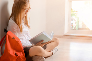 Cute schoolgirl with backpack sitting on floor and reading book indoors