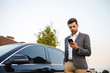 © Drobot Dean - Portrait of rich businessman wearing suit, standing near his luxury black car, and using smartphone while holding in hand