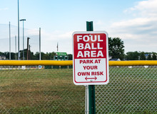 Sign At Children's Baseball Field, Warning Foul Ball Area.  Red And White Sign Warns Of Foul Balls, Park At Your Own Risk. 