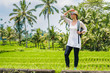 © Alexander - Beautiful young girl traveler in hat, glasses, with backpack on a rice field, in high green grass, with palms, amazing blue sky with white clouds on the background has journey in Bali island Indonesia