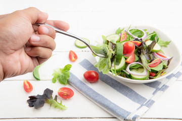 hand is using a fork to slice salad with fresh lettuce, tomatoes and cucumber in white plate on white table