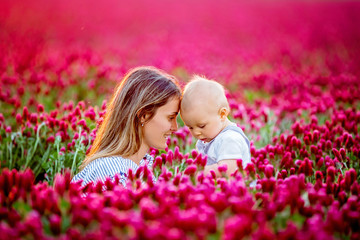 Poster - Young mother, embracing with tenderness and care her toddler baby boy in crimson clover field, smiling happily