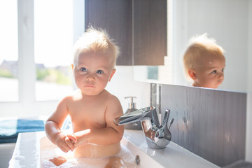 Poster - Baby taking bath in sink. Child playing with foam and soap bubbles in sunny bathroom with window. Little boy bathing. Water fun for kids. Hygiene and skin care for children