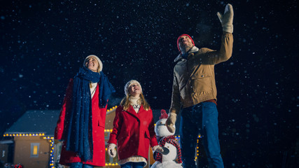 Wall Mural - Happy New Year Celebration, Young Family of Three Standing in the Front Yard Looking and Pointing into the Sky. In the Evening while Snow is Falling Father, Mother and Cute Little Daughter Look up.