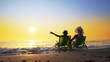 © zefart - Mother and son with hat sitting on beach chairs looking at the setting sun splashed by sea waves. Travel concept