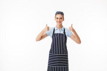 Wall Mural - Young smiling woman cook in striped apron and cap happily looking in camera while showing big thumbs up gesture over white background