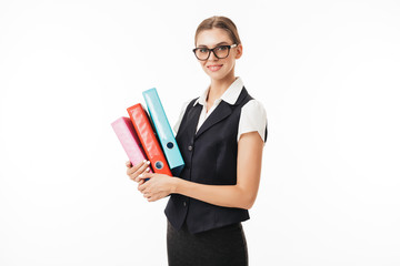 Wall Mural - Young beautiful woman in black vest and eyeglasses happily looking in camera while holding colorful folders in hands over white background