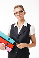 Wall Mural - Young serious woman in black vest and eyeglasses thoughtfully looking in camera while holding colorful folders and pencil in hands over white background