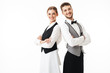 Young smiling waiter and waitress in white shirts and vests sstanding back to back while joyfully looking in camera with arms folded over white background