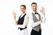 Young smiling waiter and waitress in white shirts and vests sstanding back to back happily looking in camera while showing ok gestures over white background