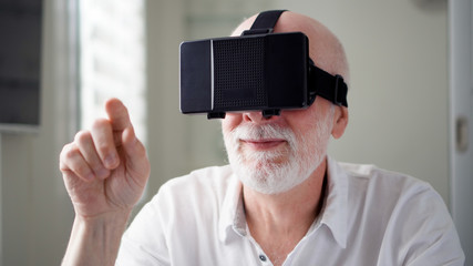 Good-looking handsome senior man in white using VR 360 glasses at home. Making browse and tap gestures. Concept of active modern elderly people, learning to use contemporary technologies.