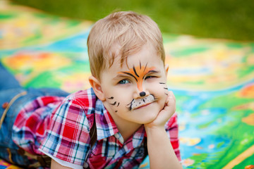 Cute little blonde boy with tiger make-up lying on the green grass