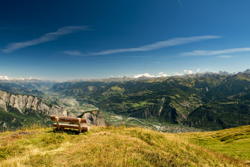 Scenery alpine panorama of Switzerland in sunny day with view on Ovronnaz, mountains, green forest, meadows, blue sky and white clouds with a bench for rest in the foreground