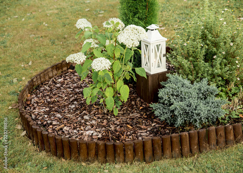 Fotomural Round flower bed covered by pine bark mulch and surrounded by brown weatherproof wood roll boarder palisade, inside are different plants, outdoors in summer