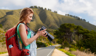 Wall Mural - travel, tourism and photography concept - happy young woman with backpack and camera photographing over big sur hills and road of california background