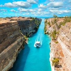 Beautiful scenery of the Corinth Canal in a bright sunny day against a blue sky with white clouds. Among the rocks floating white ship in turquoise water.