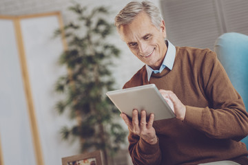 Modern technologies. Pleased senior man keeping smile on his face while looking at screen of his gadget